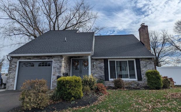 Stone-faced ranch house with a gray garage door, black shutters, and brick chimney on a cloudy day.