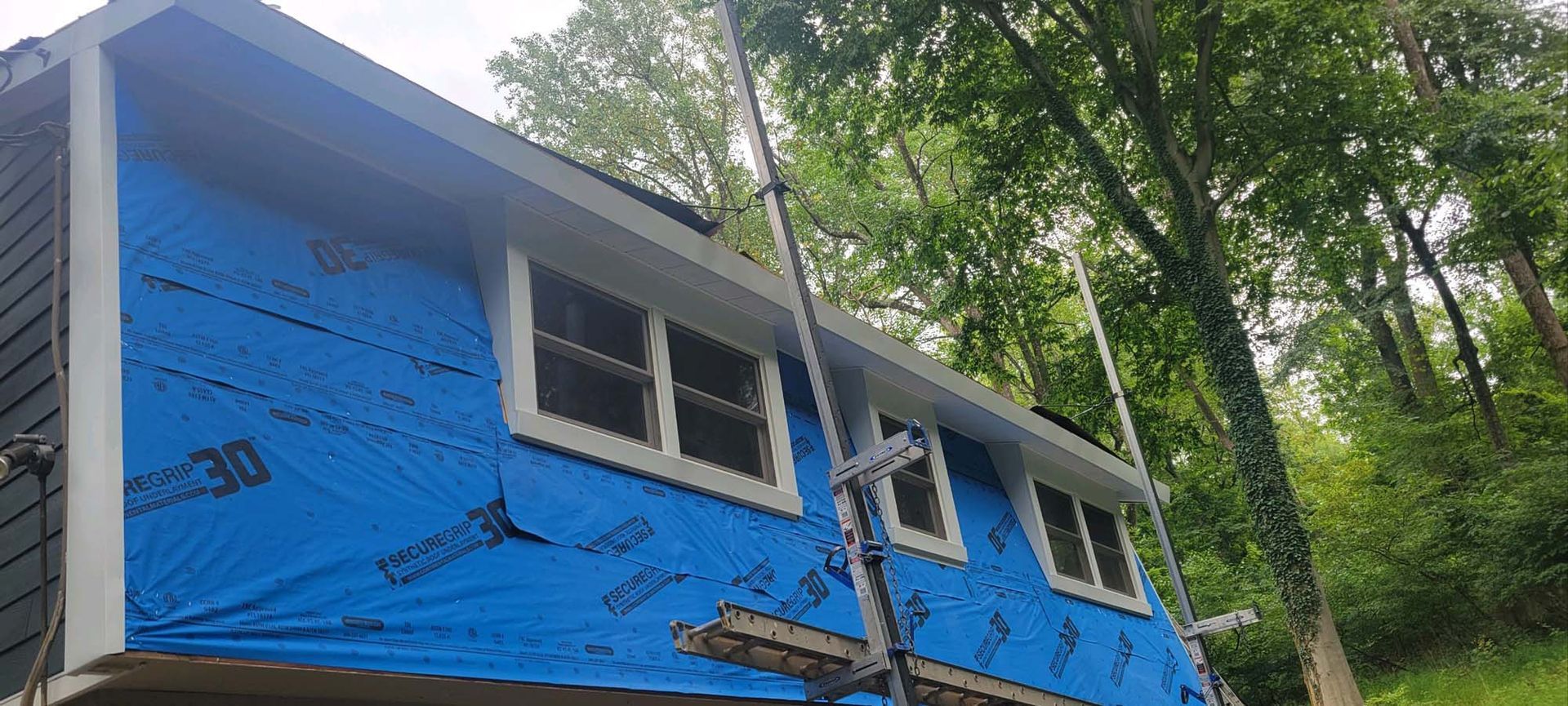 House siding under construction, blue wrap, white trim, scaffolding, trees in background.