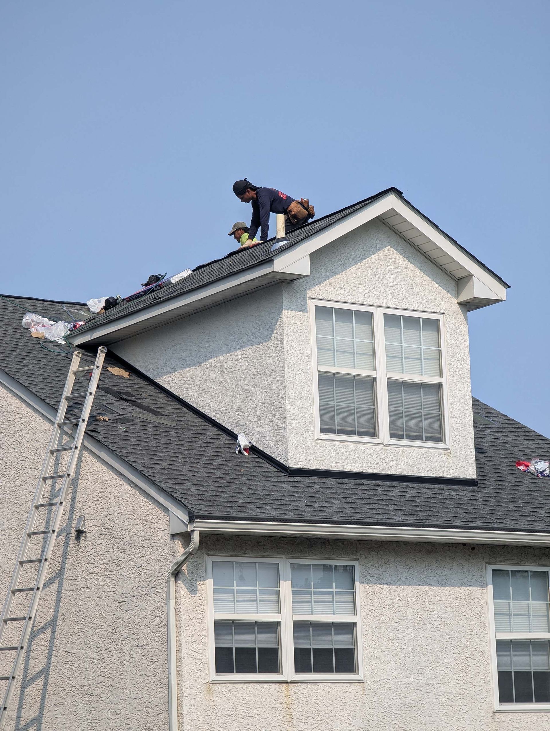 Roofer working on a house roof. Ladder, blue sky, and stucco siding are visible.