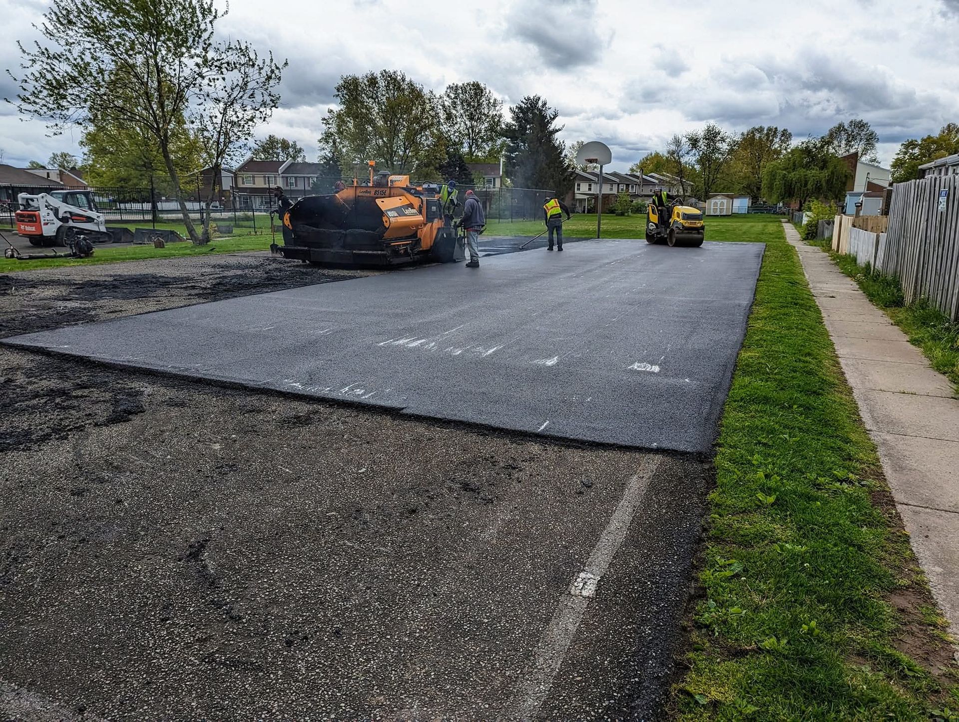 Paving crew laying asphalt on a road. Equipment includes a paver and roller. Green grass, gray sky.
