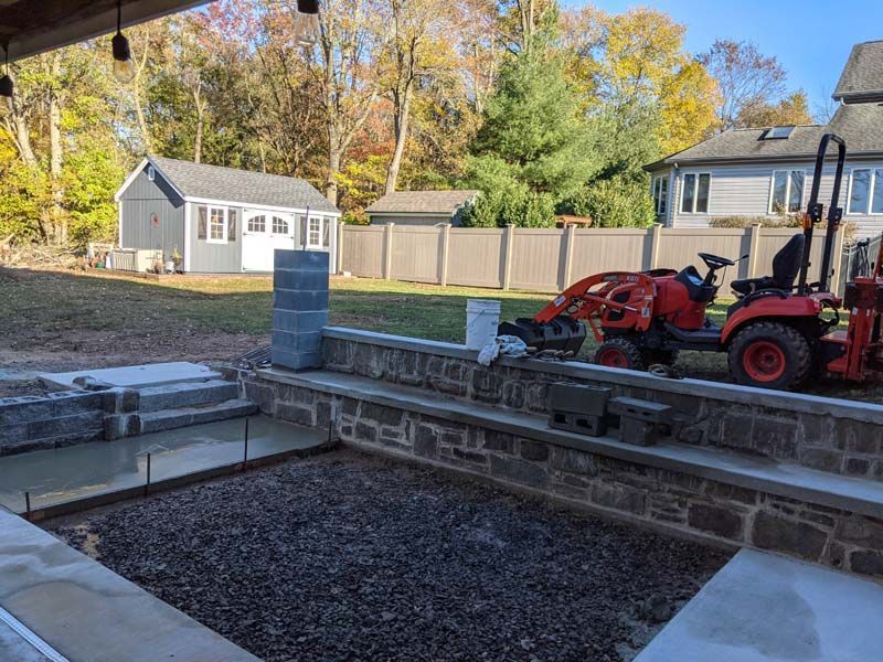 Construction site with a tractor, stone retaining wall, and concrete patio being built in a backyard.
