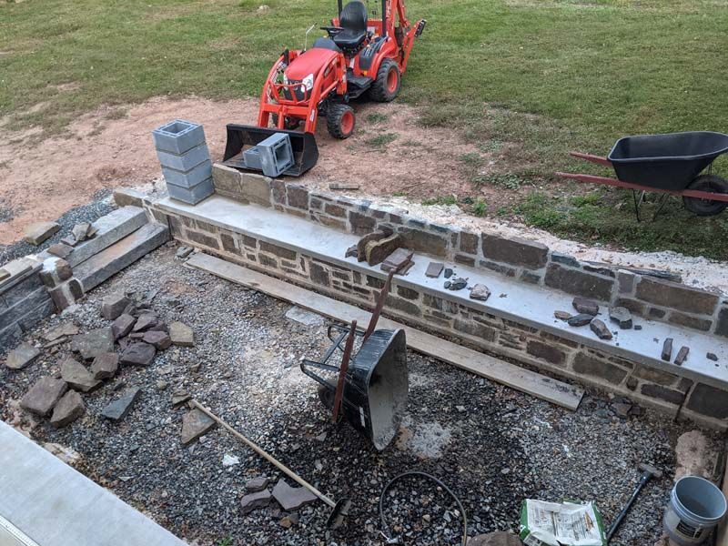 Construction site: Stone walls, a tractor, wheelbarrows, and tools on dirt and gravel.