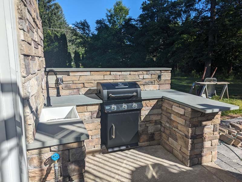 Outdoor kitchen with stone masonry, built-in grill, sink, and countertop. Blue sky background.