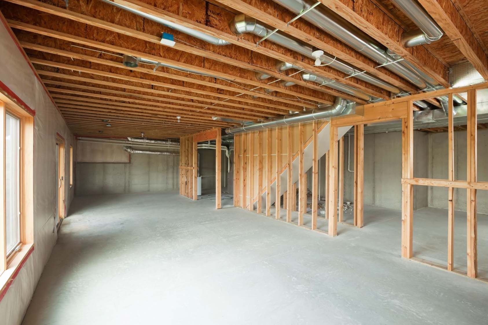 Unfinished basement with wooden framework, concrete floor, windows, and exposed pipes and ceiling beams.