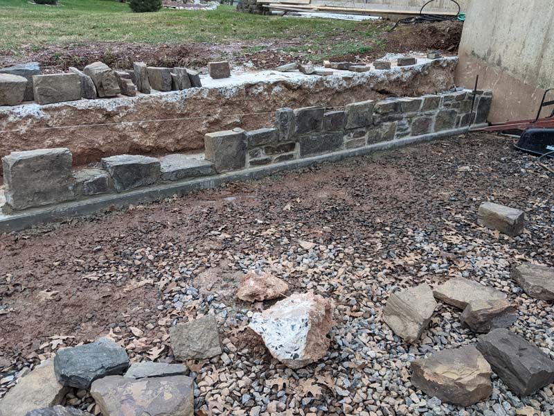 Construction of a low stone wall, with foundation visible. Rocks and soil surround the partially built structure.