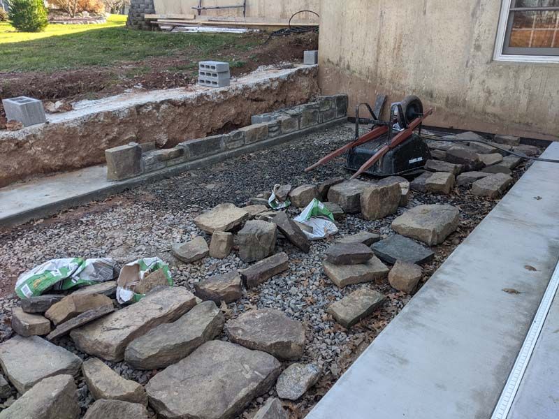 Construction site with stones, gravel, wheelbarrow, and foundation work near a building.