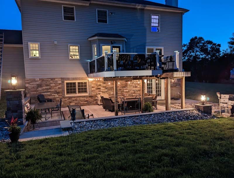 Backyard at dusk with deck, patio, and illuminated landscaping against a two-story house.