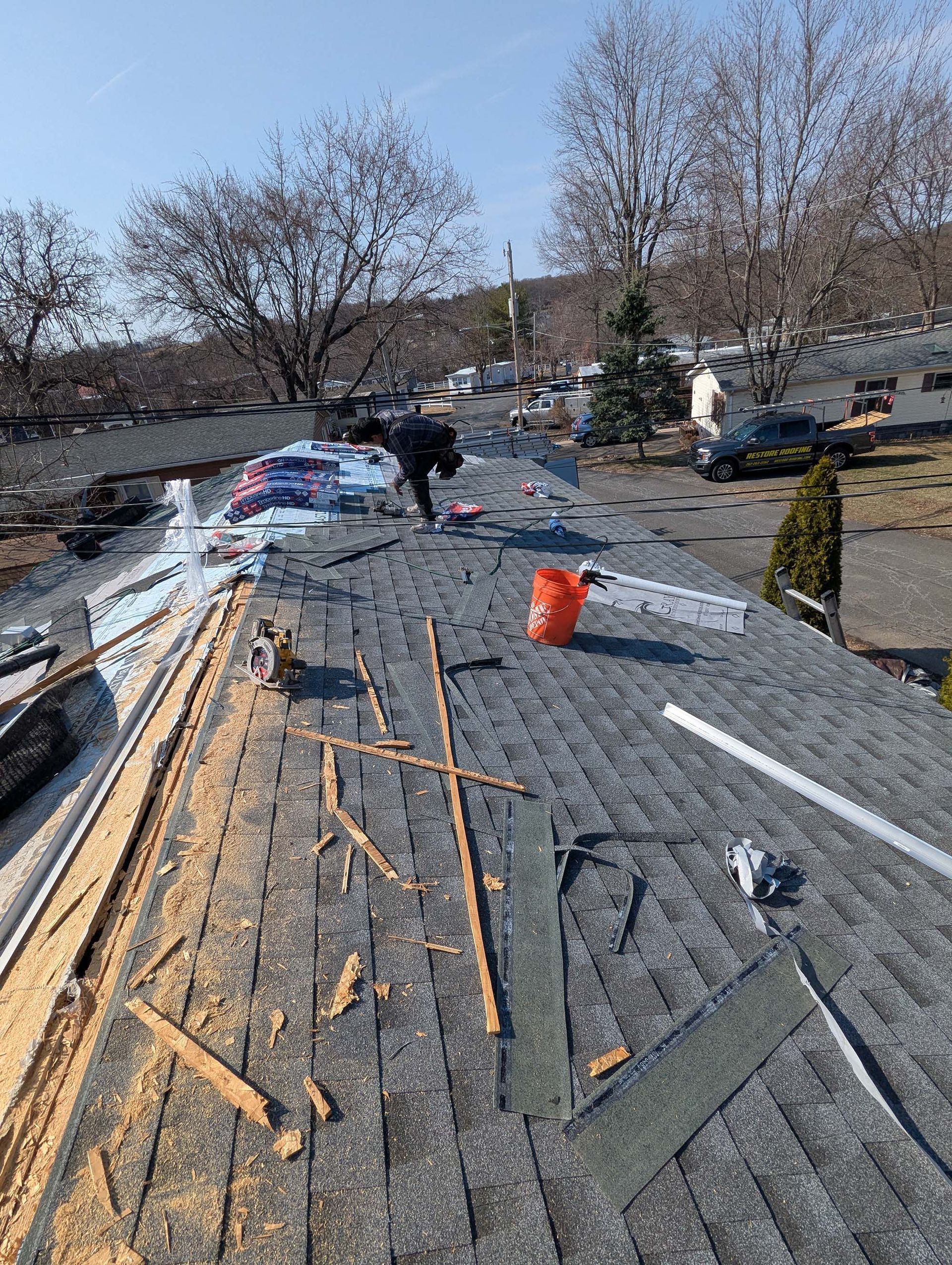 Person on a rooftop replacing shingles. Orange bucket, construction debris, clear sky.