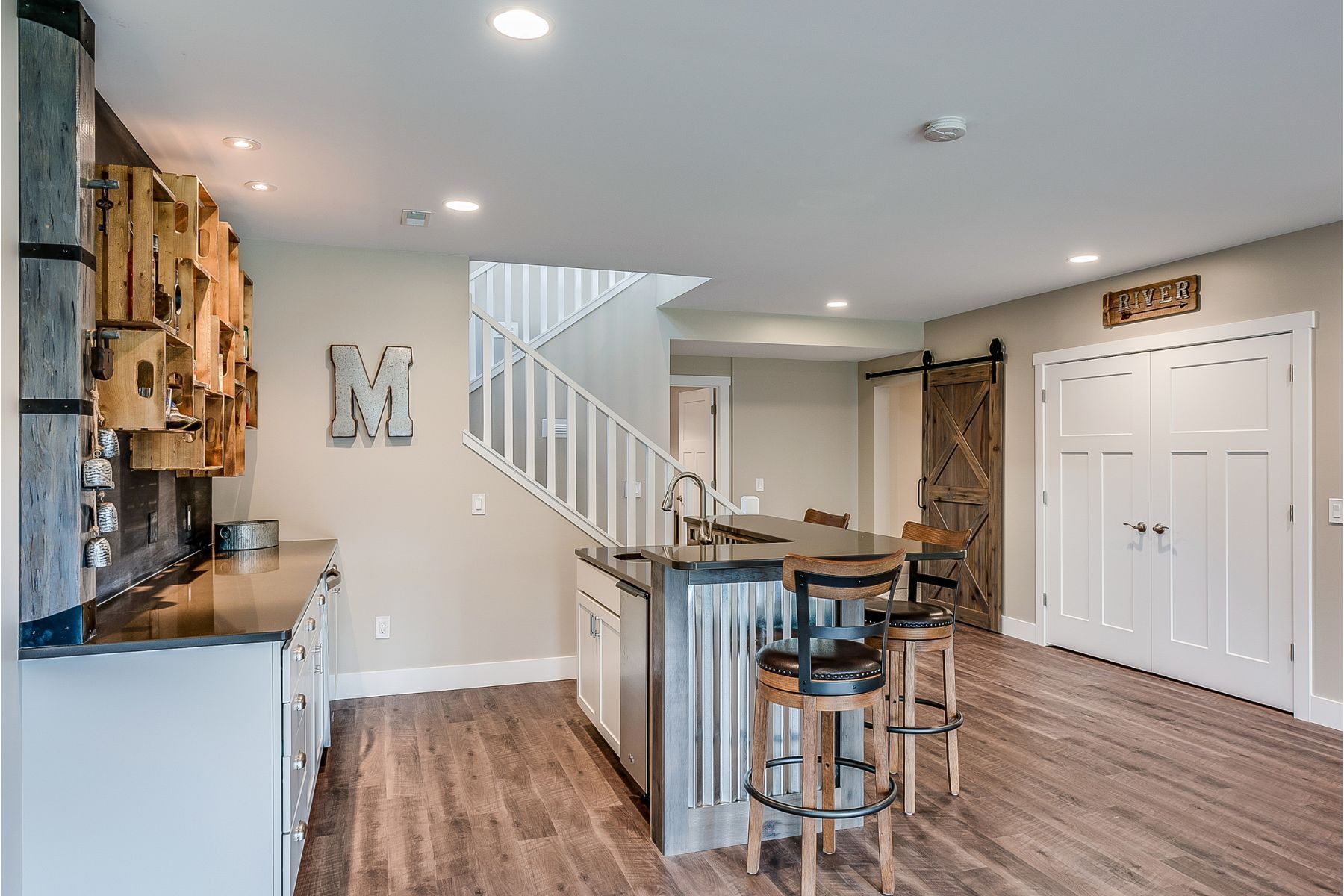 Basement with kitchen, bar, stairs, and wooden accent wall. Dark wood floors, white cabinets and walls.