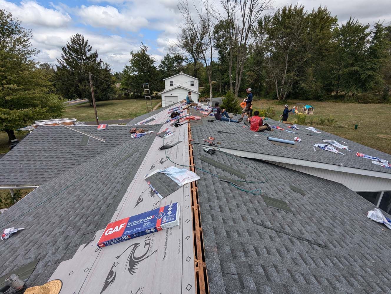 Roofers working on a house roof. Various materials and tools are visible. Green trees and sky in the background.