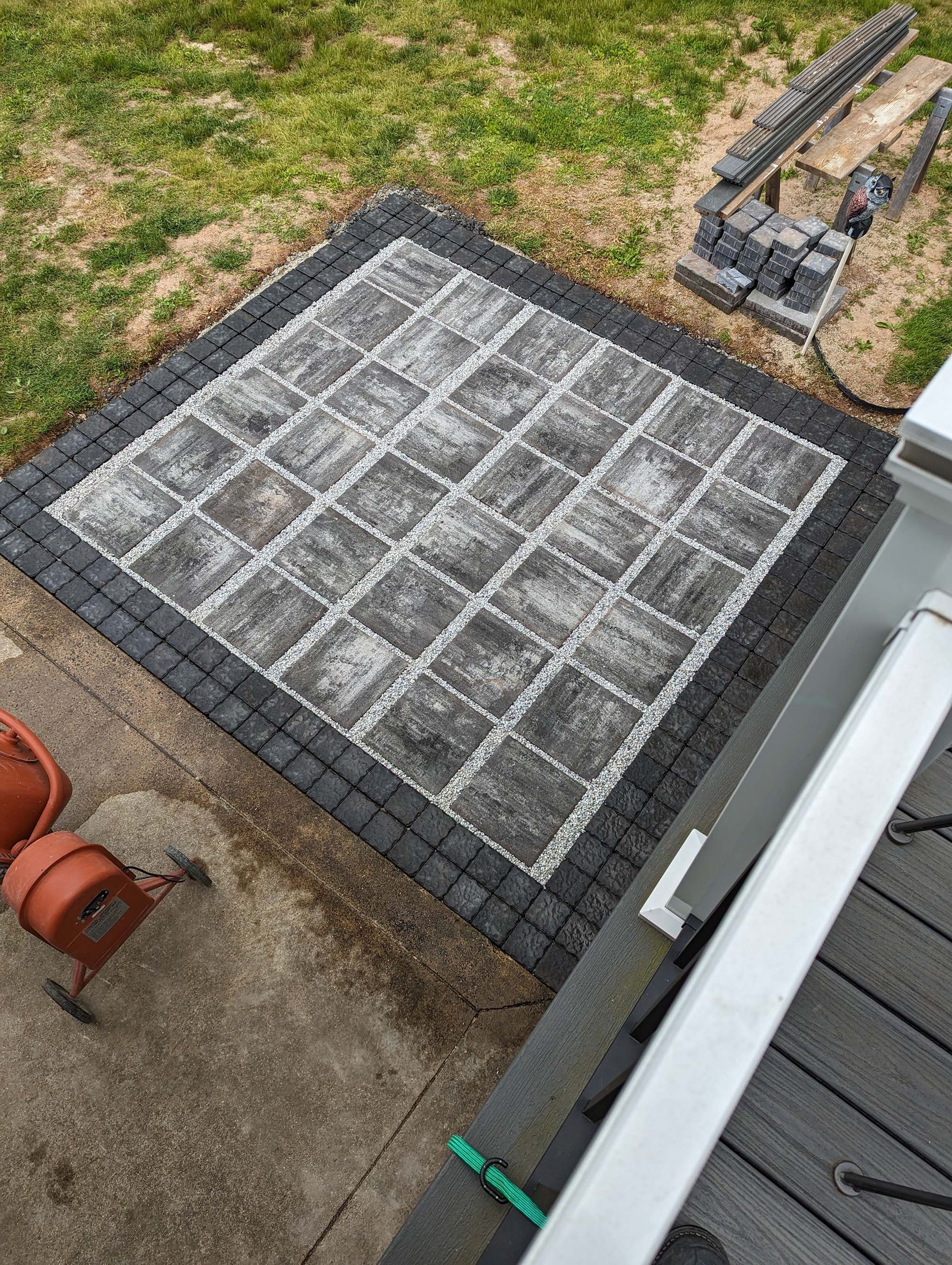Square patio with gray and black patterned pavers. Brick border surrounds the design.