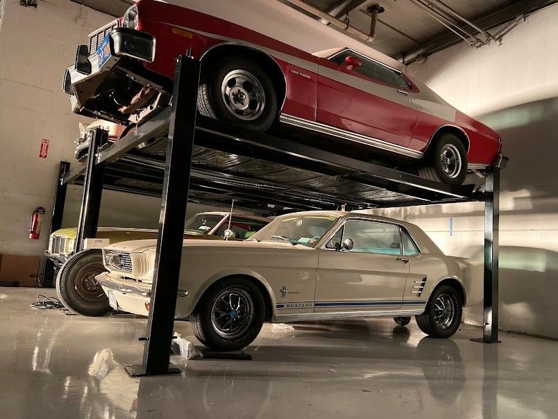 Classic cars stacked on a black lift in a garage. Red car on top, tan Mustang below.
