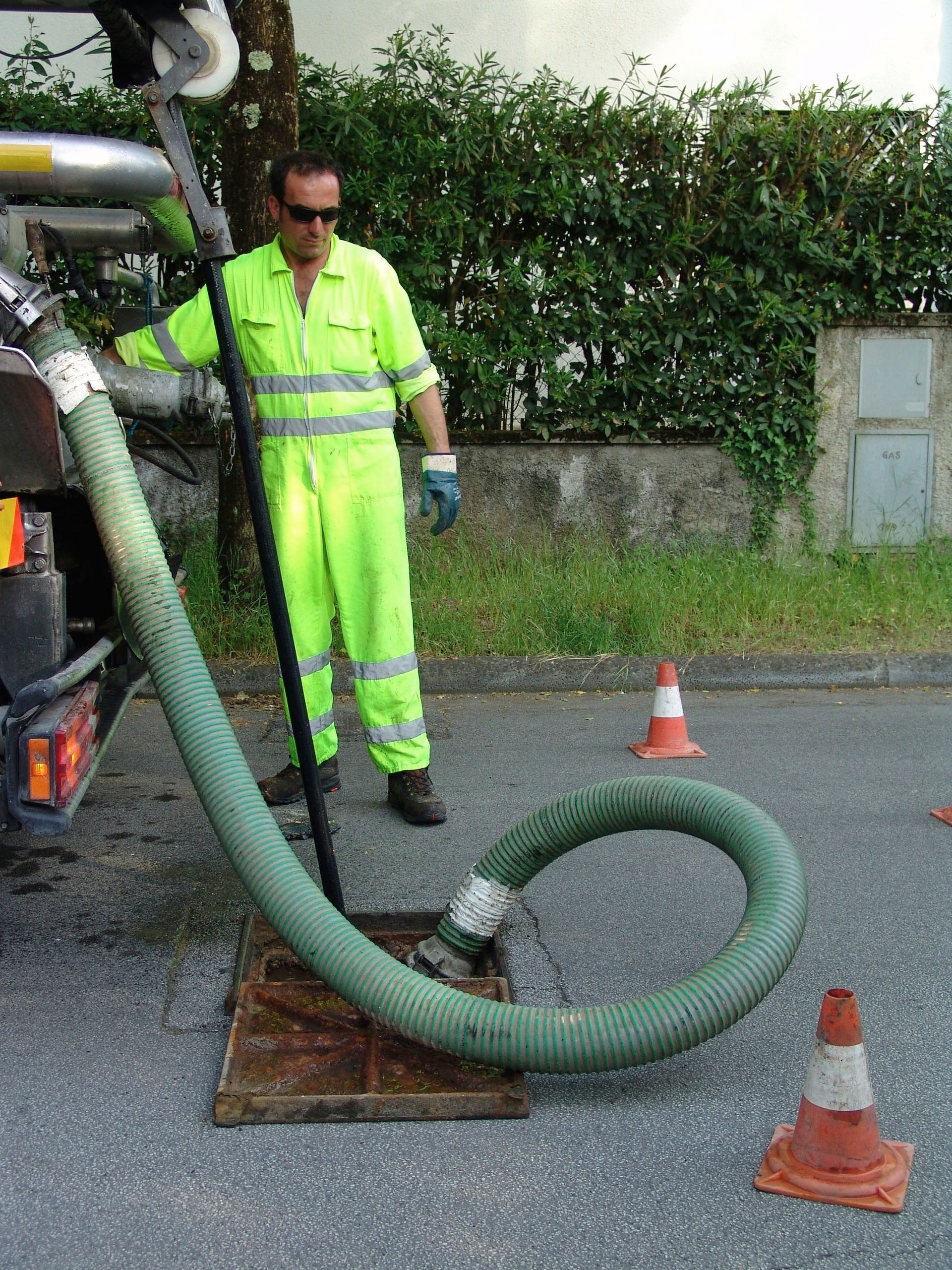 Man in neon suit working on a sewer with a hose connected to a truck. Orange traffic cones on the street.