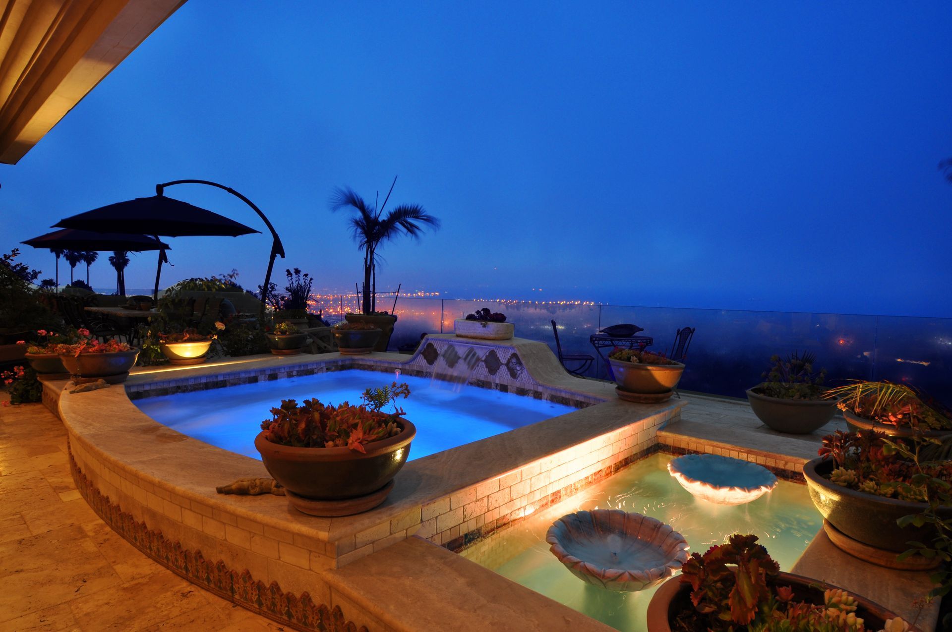 Pool area at dusk with illuminated water, potted plants, and city lights in the background.