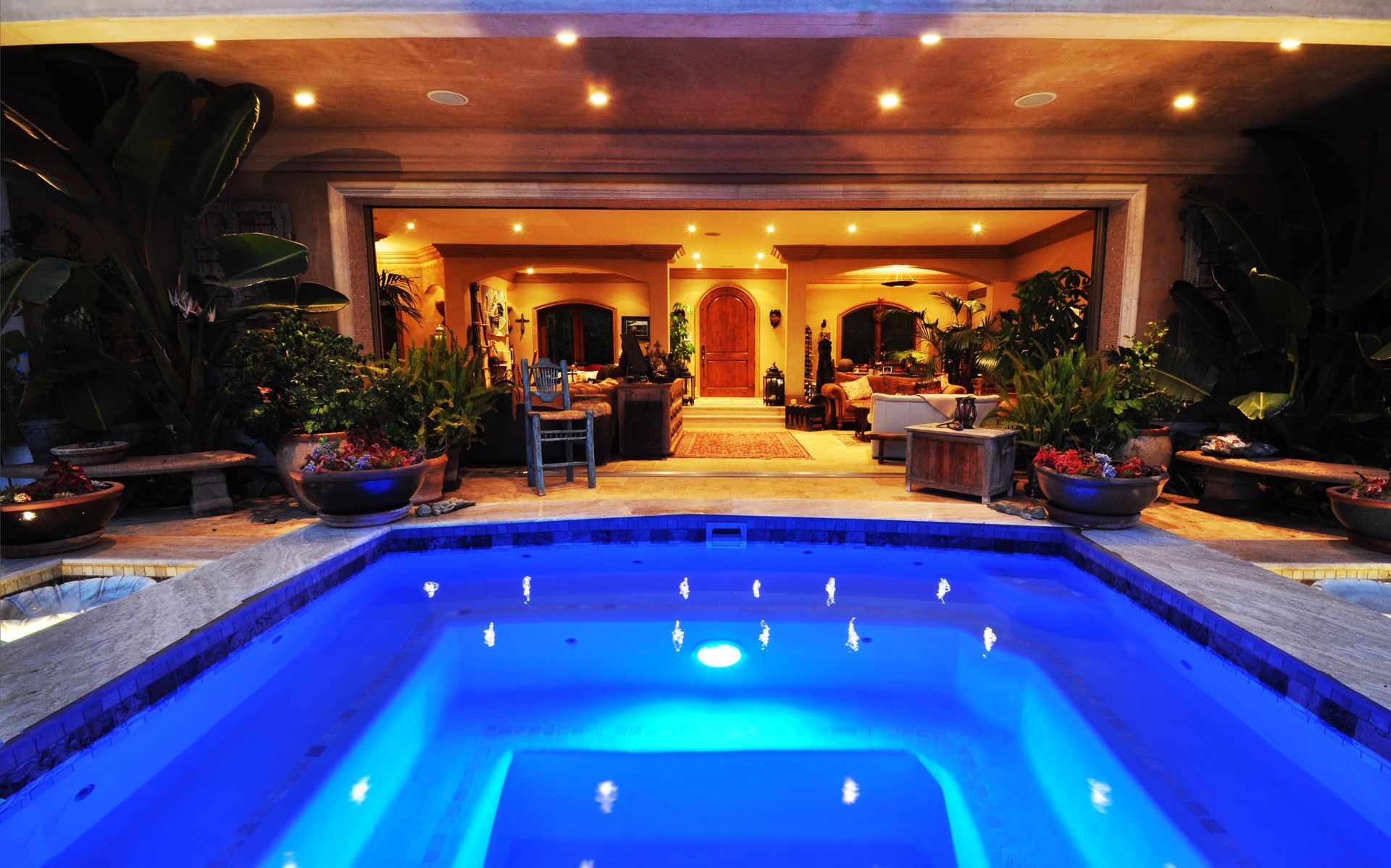 Pool with blue light, leading to an open doorway with interior lighting. Plants and seating surround the pool.
