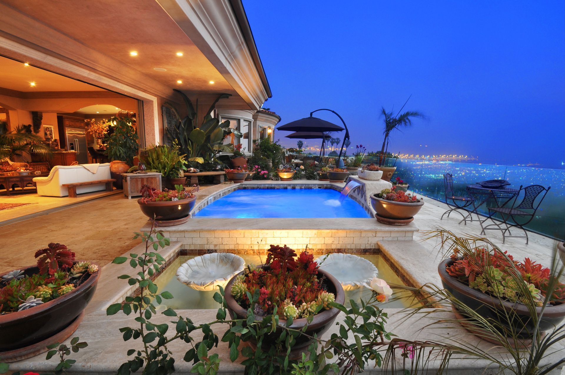 Patio with pool, flower pots, and city lights in the background at dusk.