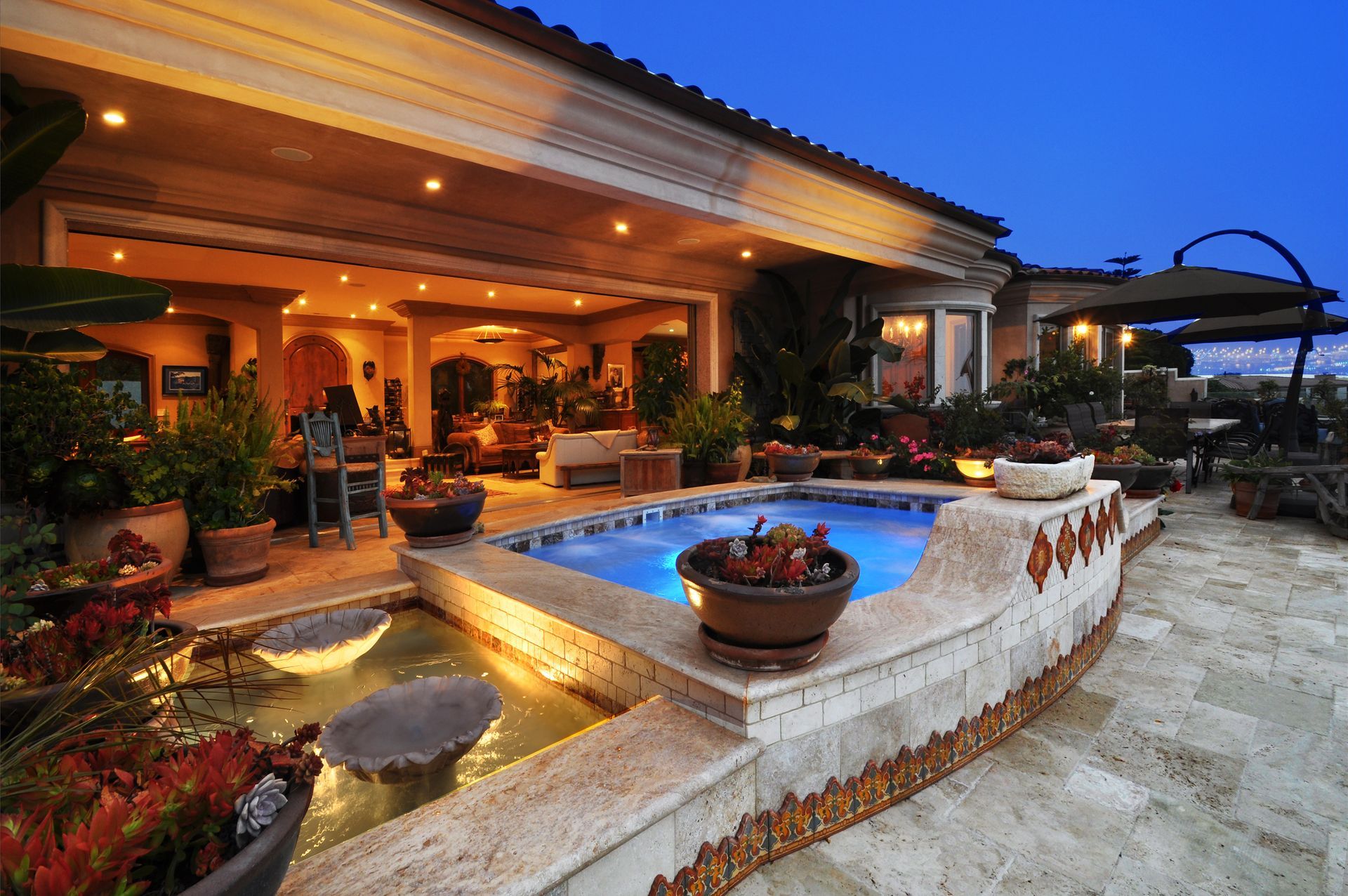 Outdoor patio with pool, fountain, and potted plants, lit up at dusk.