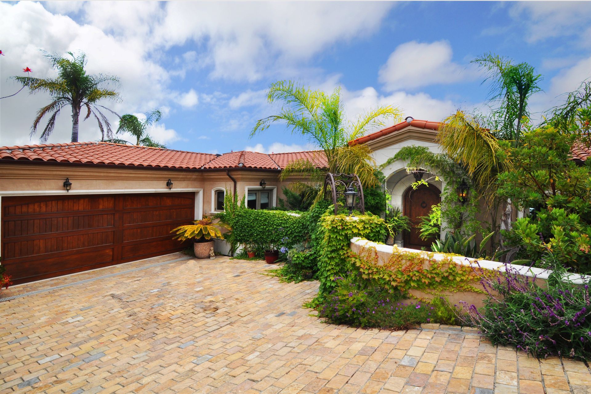 A Mediterranean-style house with a brick driveway, garage, and lush landscaping on a sunny day.