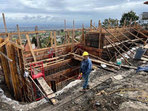 Construction worker stands near wooden concrete forms on a hillside overlooking a city. Cloudy sky.