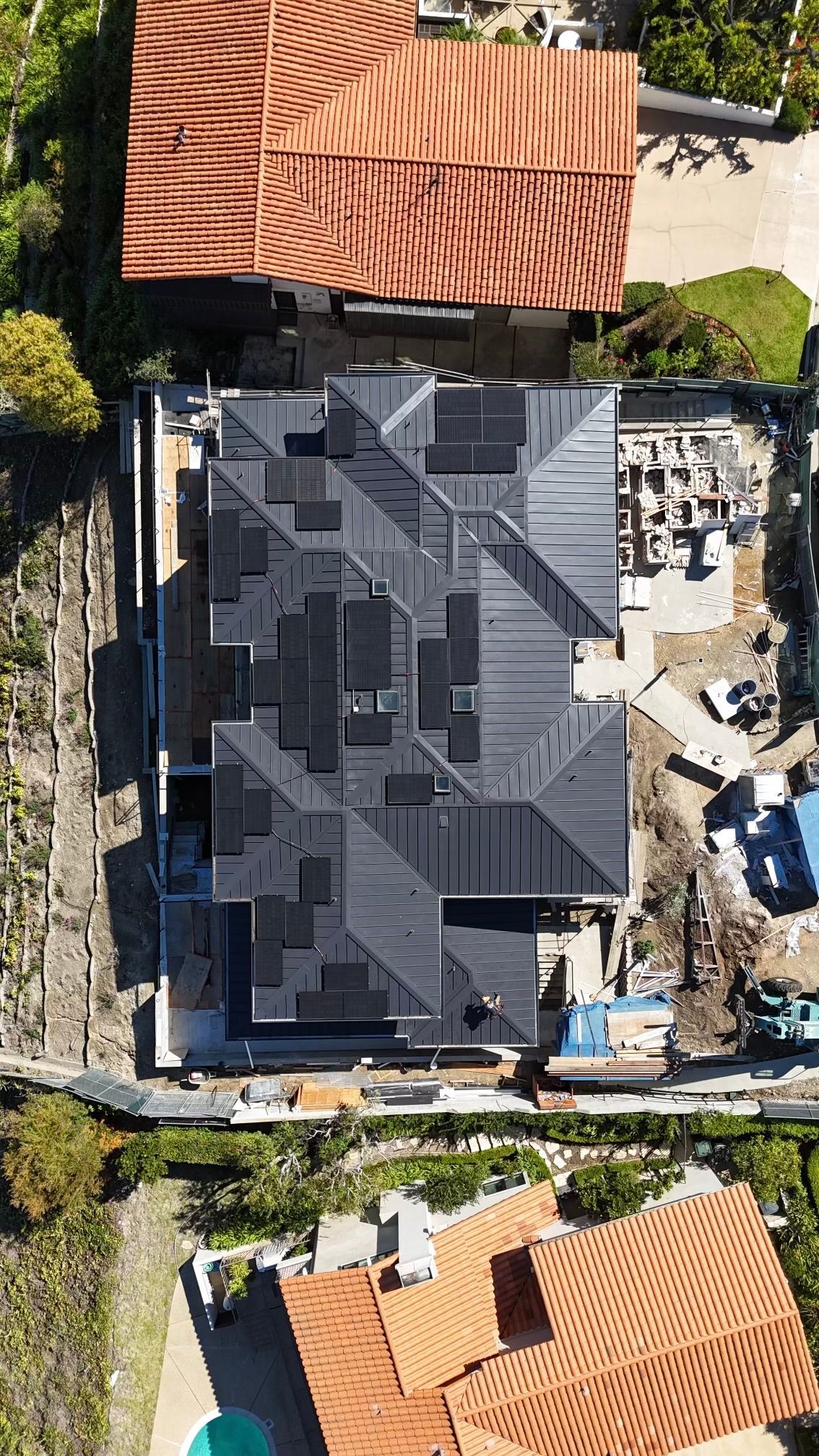 Aerial view of a dark-roofed house under construction, surrounded by other buildings with red-tiled roofs.