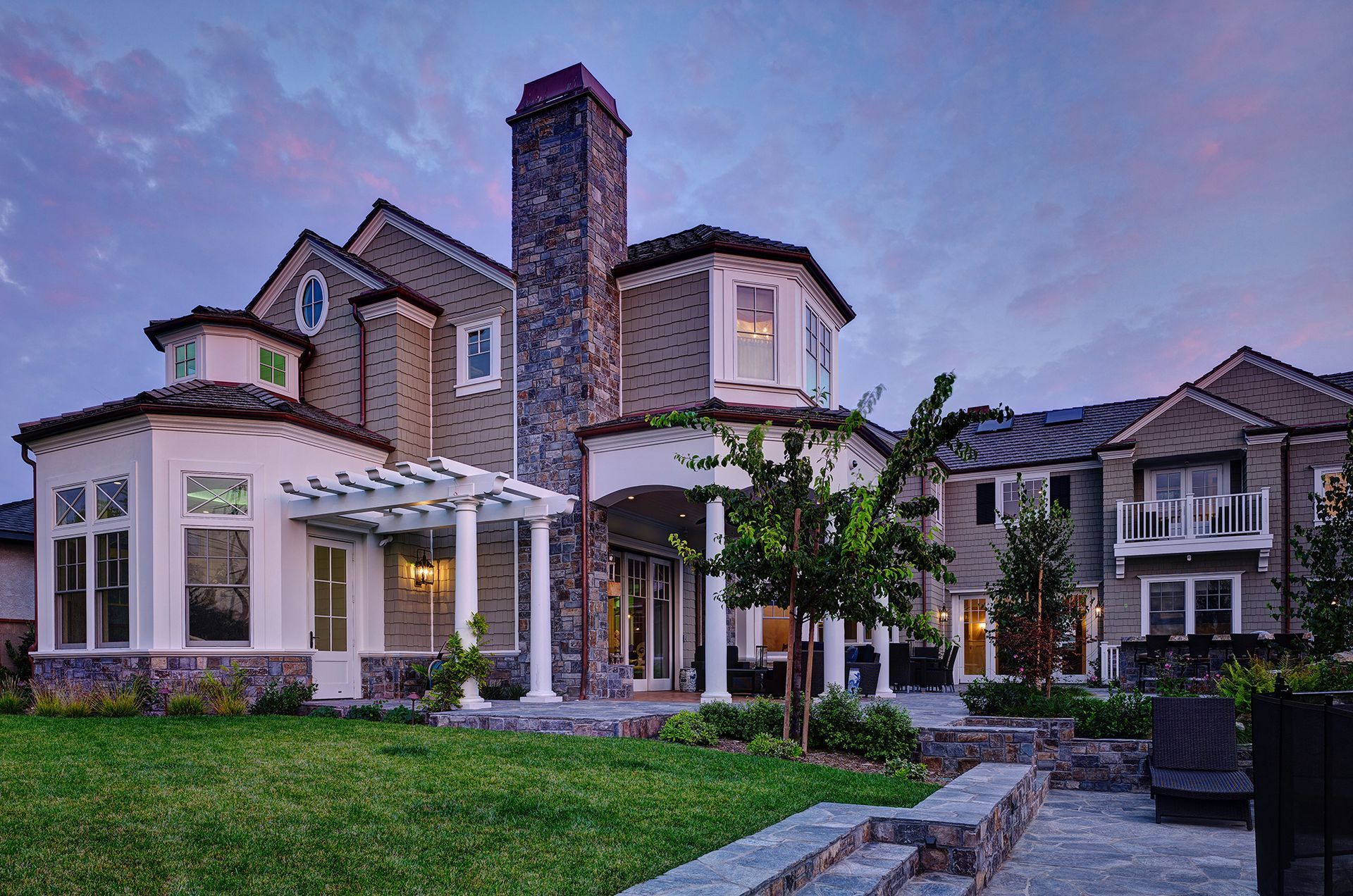 Large multi-story home with stone chimney, pergola, and manicured lawn at dusk.