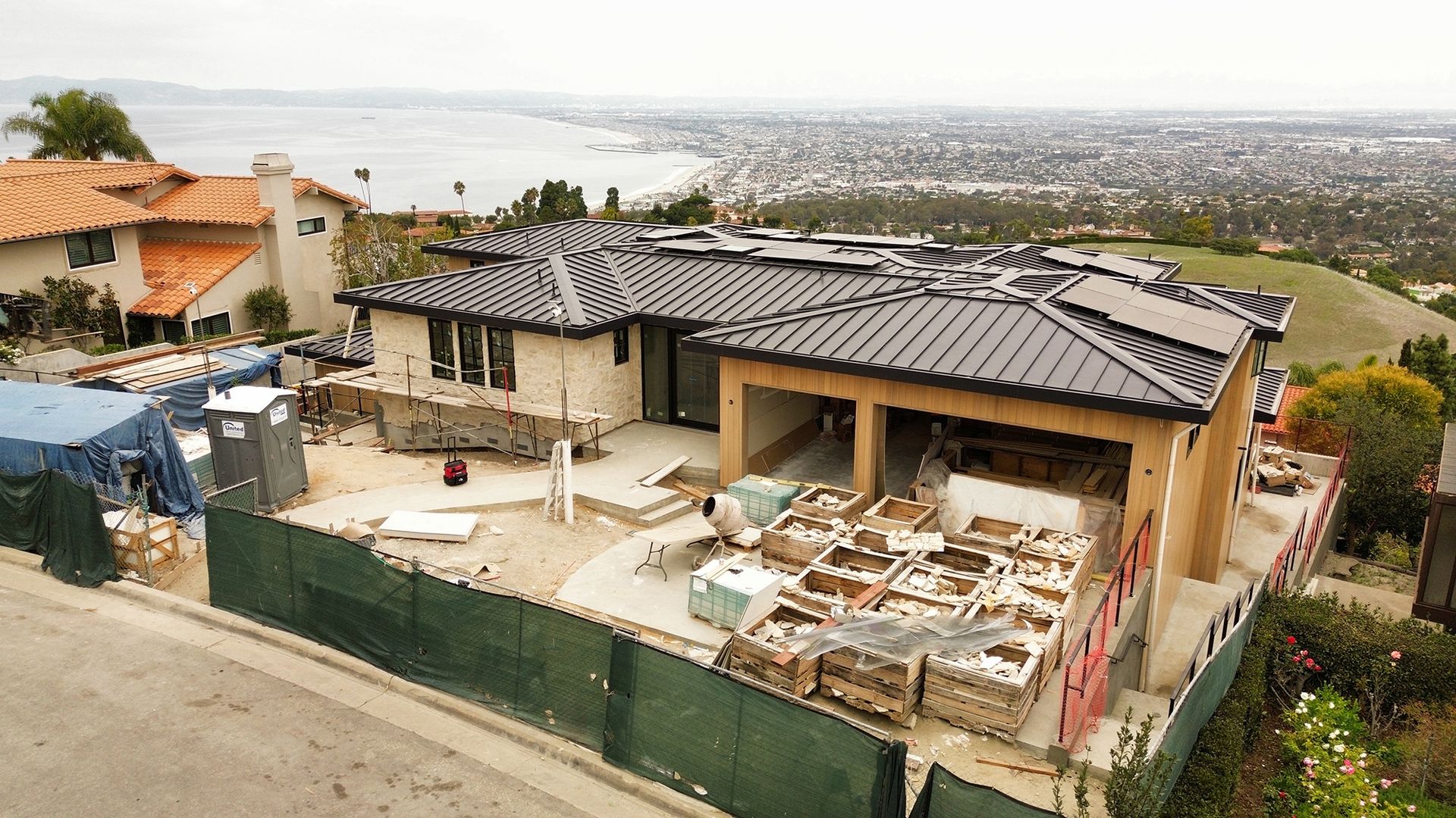 Construction site of a house with ocean view, surrounded by green fencing and materials, cloudy day.