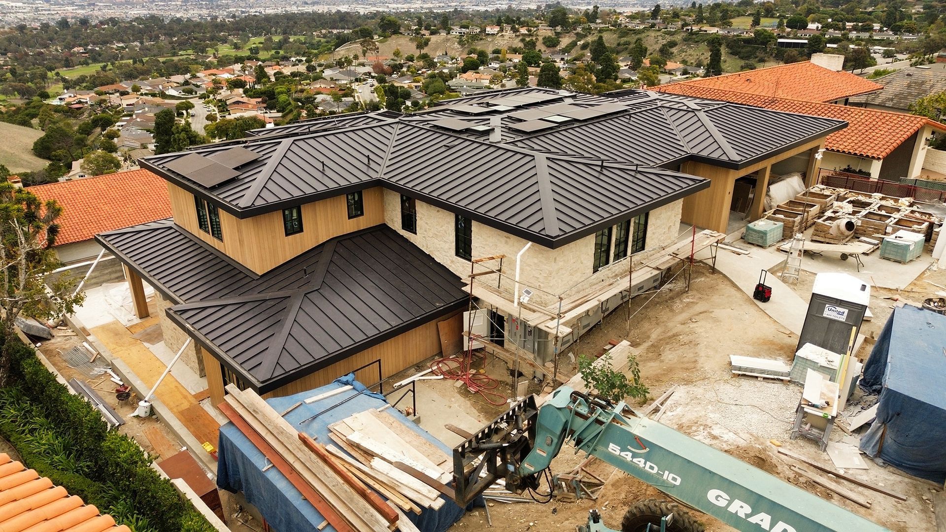 Construction site, partially built two-story house with dark gray tile roof, surrounded by other homes on a hillside.