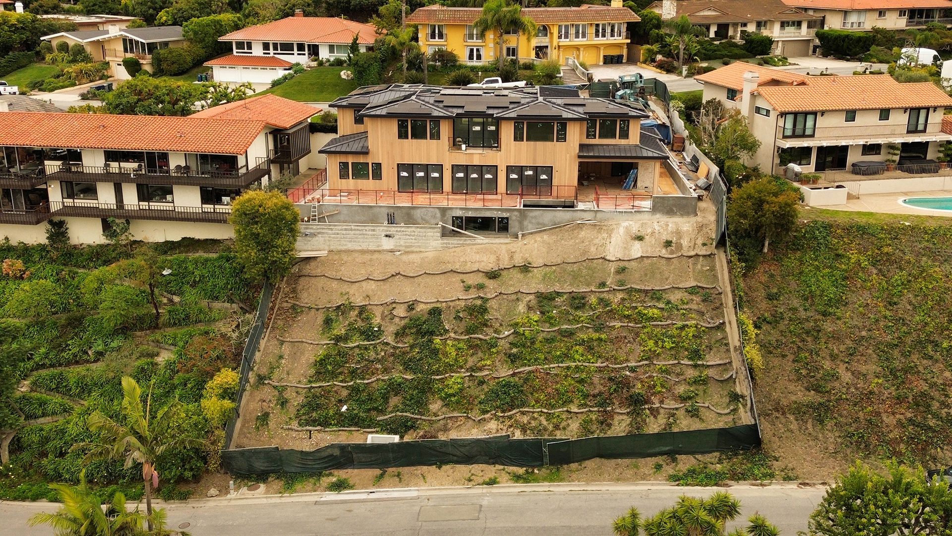 Large house under construction on a hillside, surrounded by other houses and greenery.