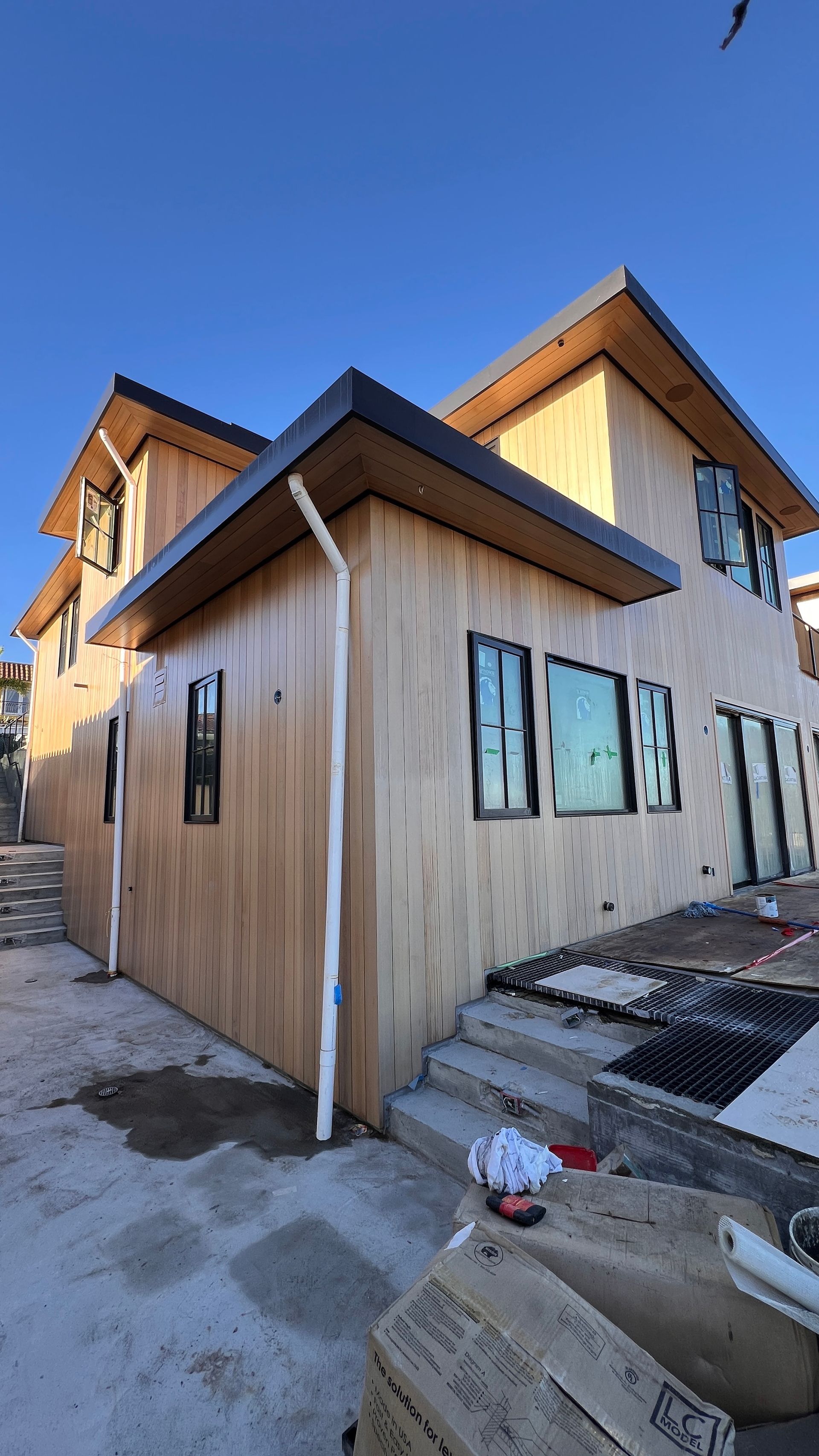 Two-story wood-clad house under construction with black window frames, dark trim, and a clear blue sky.