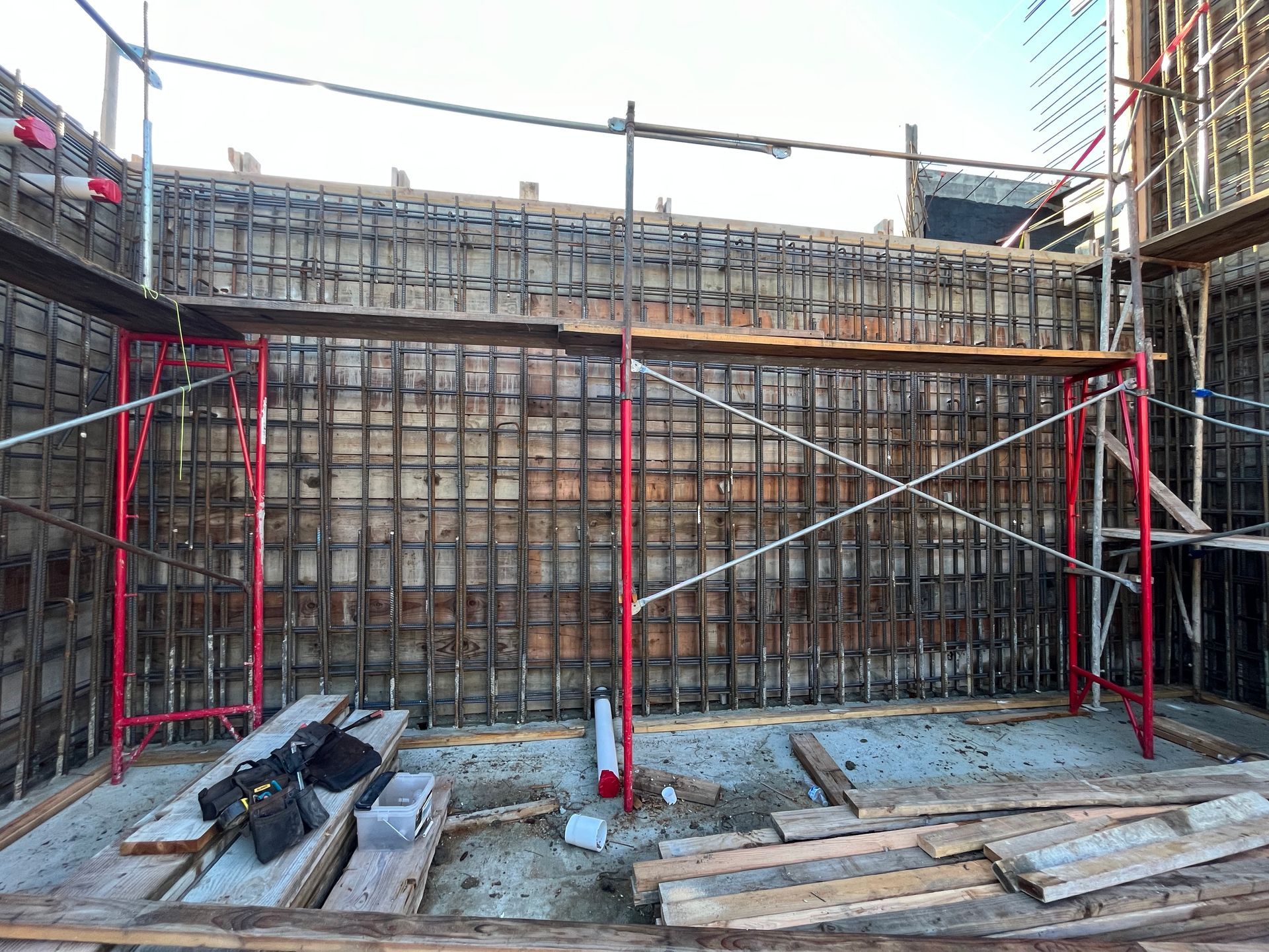 Construction site with red scaffolding, steel reinforcement, and wooden forms for concrete walls.