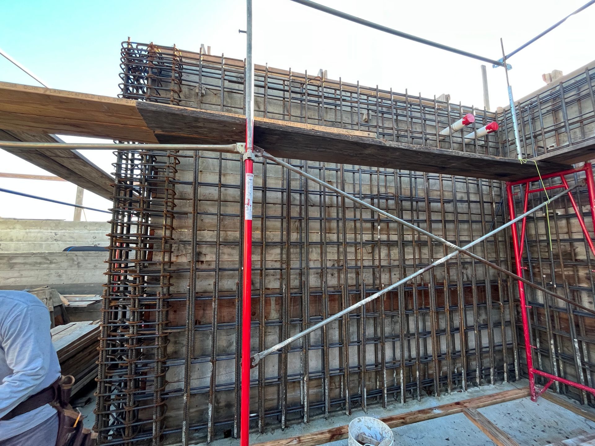 Reinforced concrete wall under construction with steel rebar framework. Scaffold and wooden planks are visible.