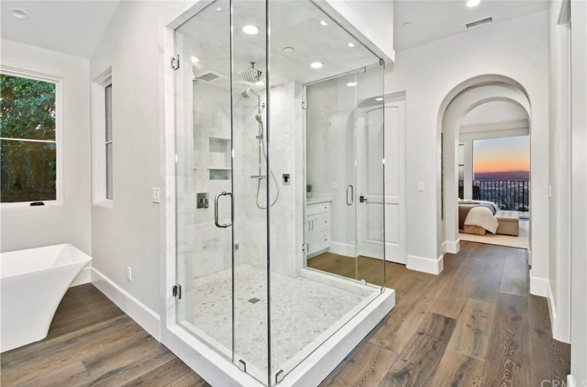 Spacious white bathroom with glass shower, wooden floor, and arched doorway to a bedroom with city view.