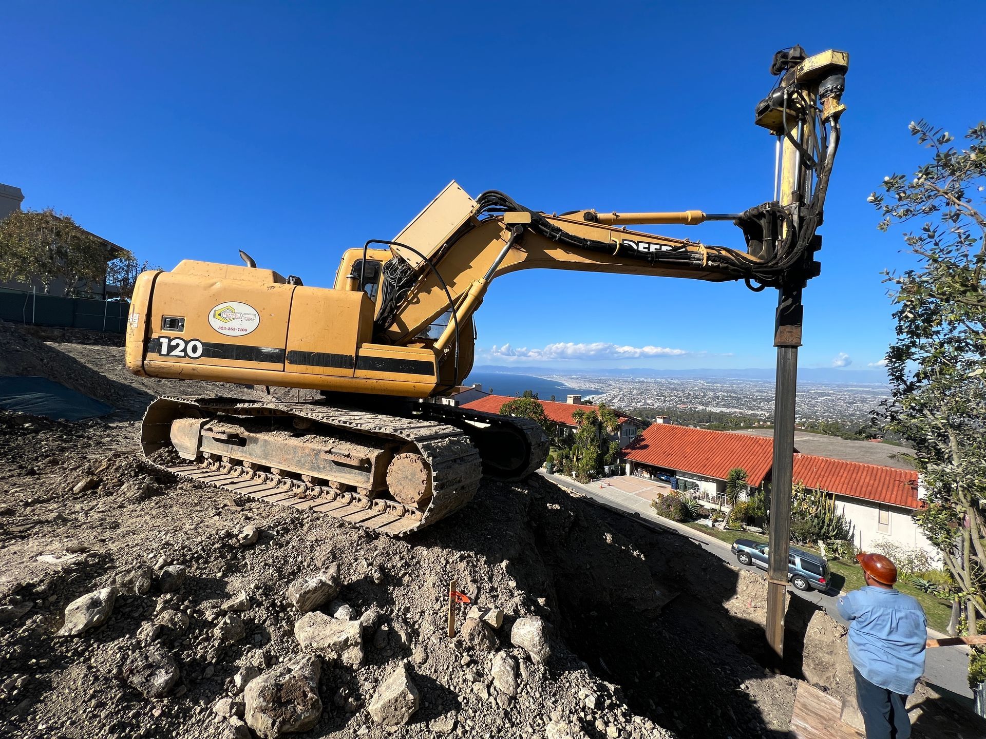 Yellow construction excavator drilling on hillside, city view in the background, clear blue sky.