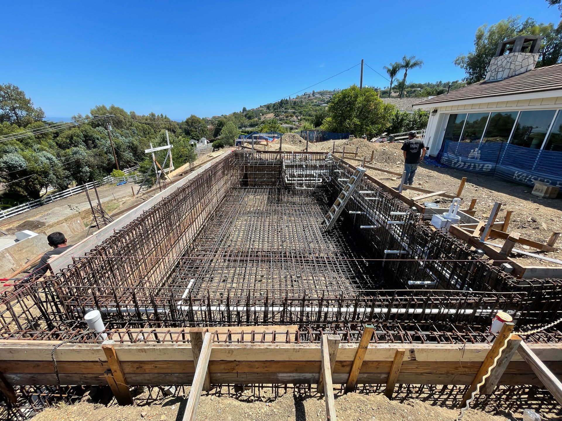 Pool construction site. Rebar framework within wooden forms; workers near a house under a bright blue sky.