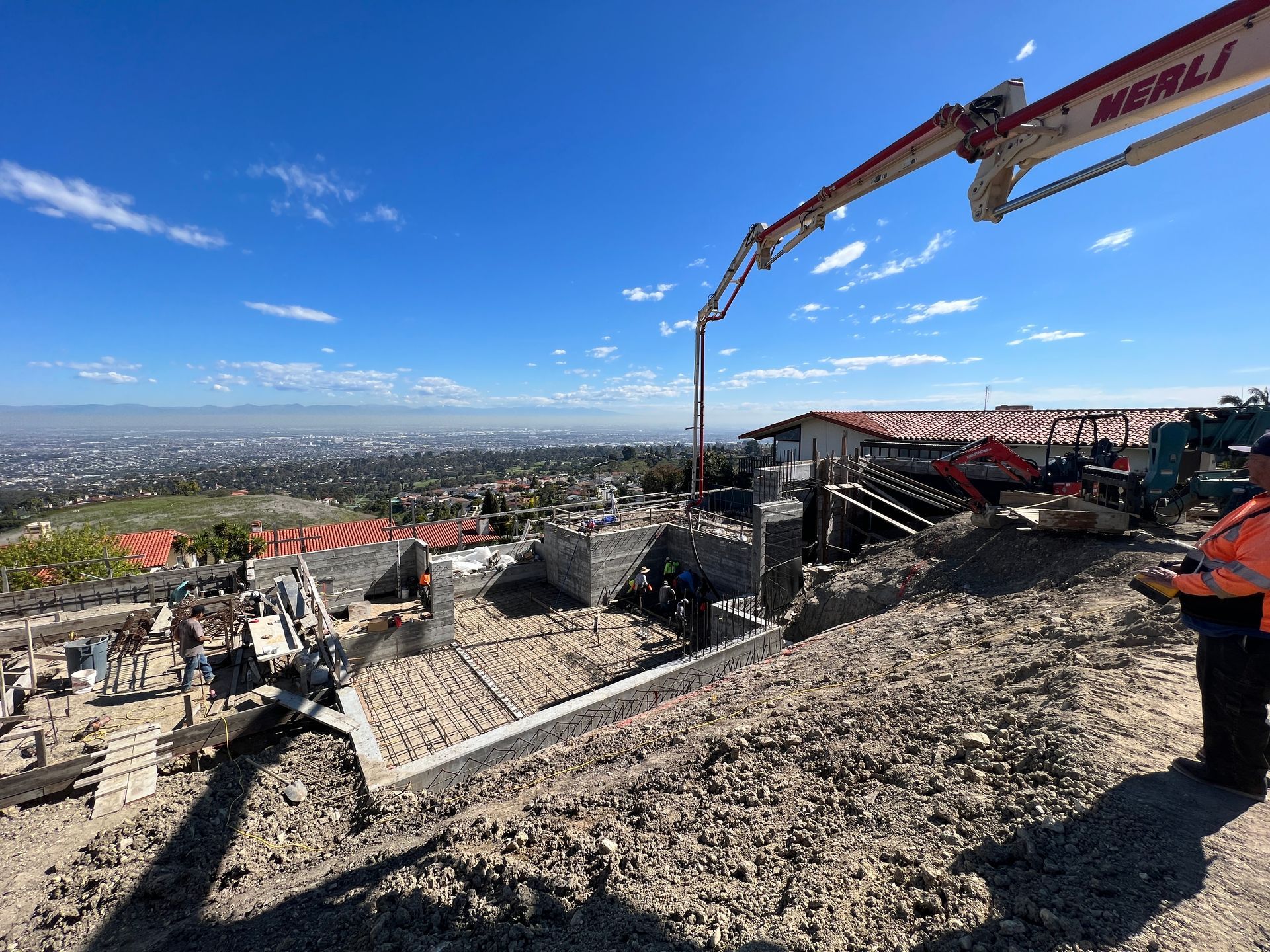 Construction site on a hillside: a concrete pump pours concrete into a building foundation, city view in the distance.