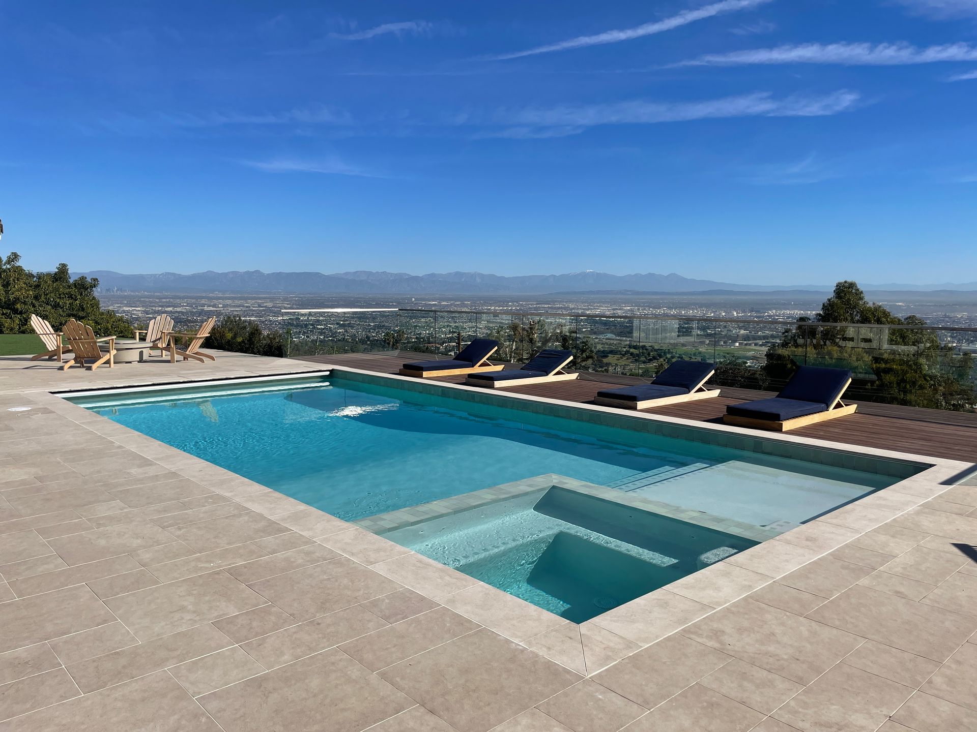 Pool with blue water and chaise lounges, overlooking a city and mountains under a blue sky.