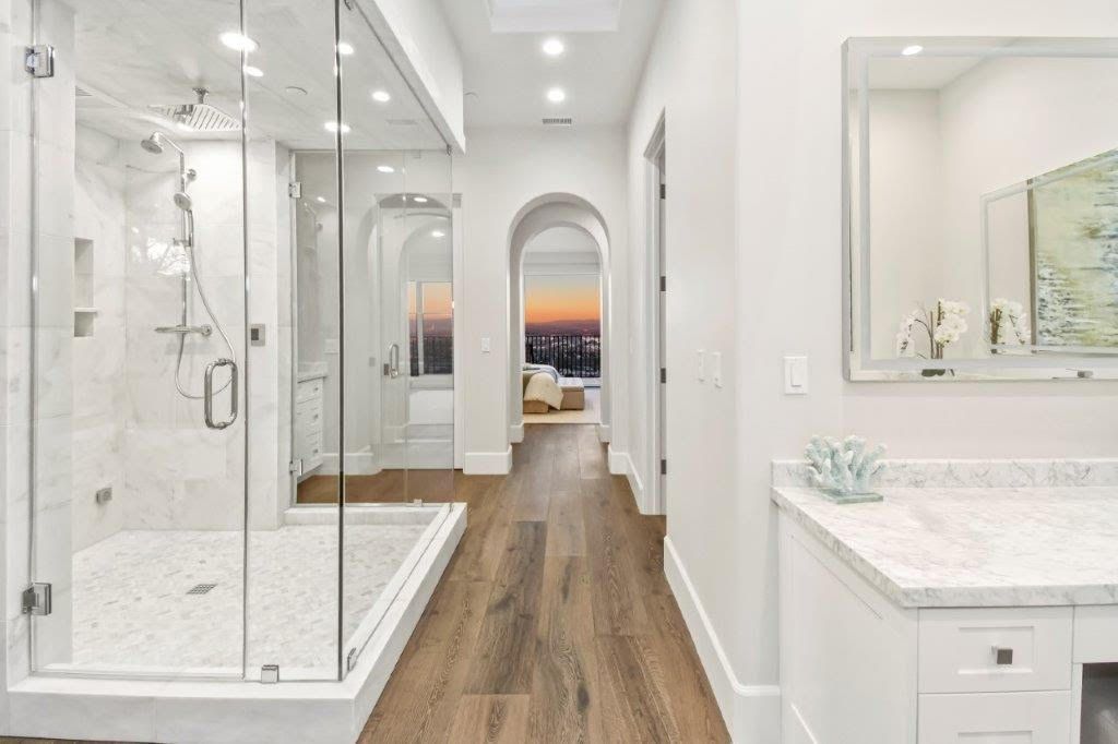 Spacious, white bathroom with wood floors. Glass shower, vanity, and arched doorway leading to a sunset view.