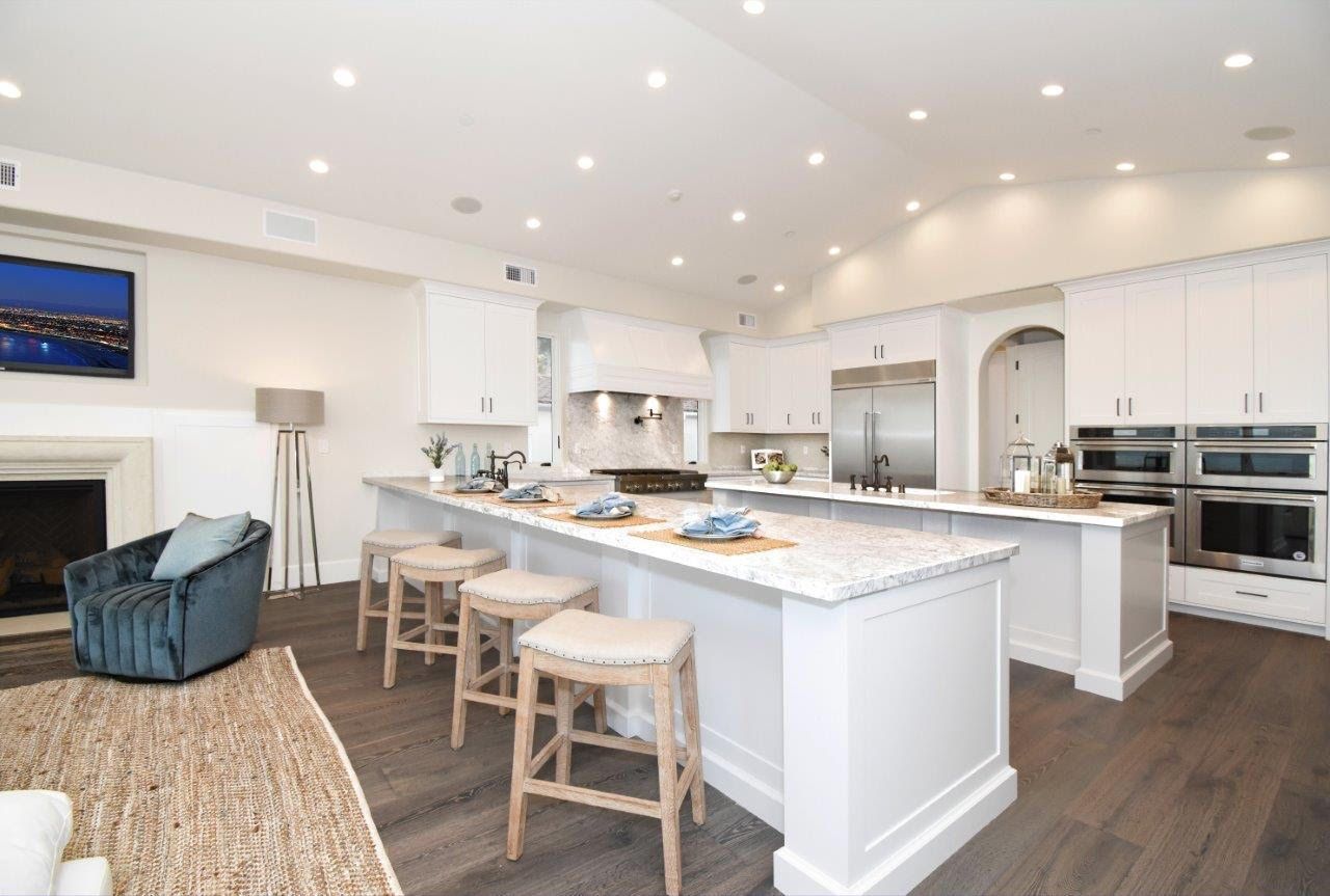 Bright, spacious white kitchen with a large island, stools, stainless steel appliances, and a connected living area.