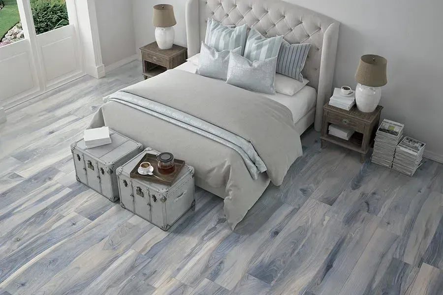 Bedroom with blue-toned wood-look flooring, light gray bed, and matching nightstands. Sunlight streams in from a doorway.