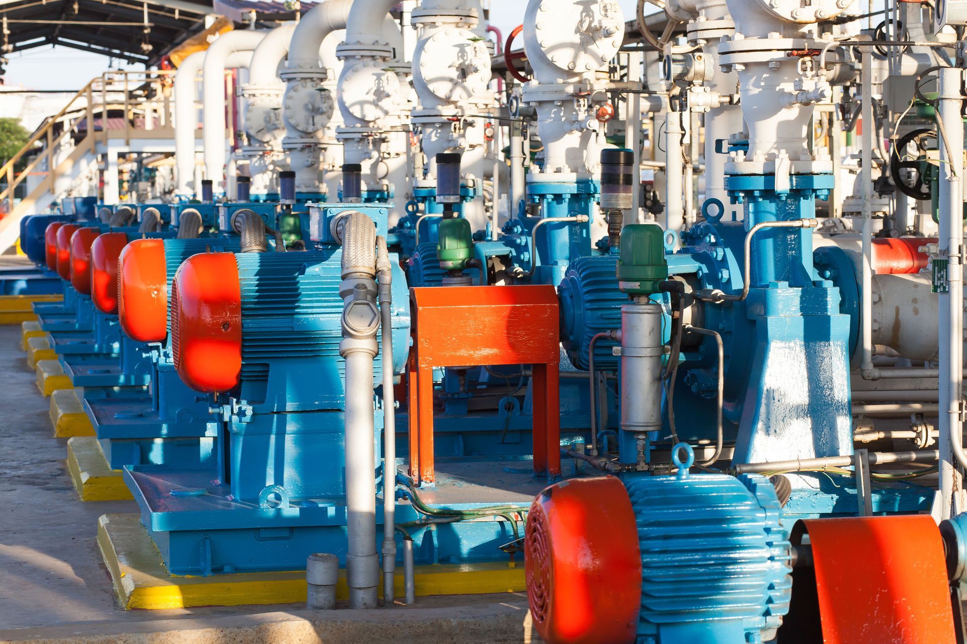 A row of blue industrial pumps with bright orange motor housings and white piping in an outdoor facility.