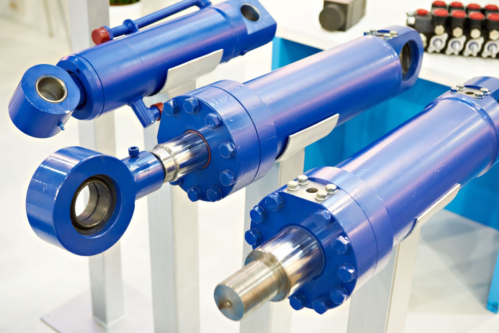 Three blue hydraulic cylinders displayed on white stands, highlighting their metal rods and mechanical joints.