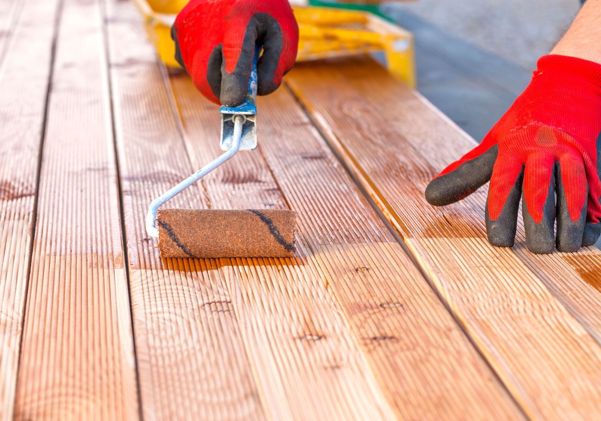Person in red gloves uses a roller to stain wood planks outside.