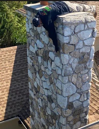 Person lying face-down on a stone chimney atop a shingled roof.