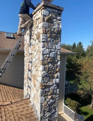 Worker on a ladder repairing a tall stone chimney on a house roof under a blue sky
