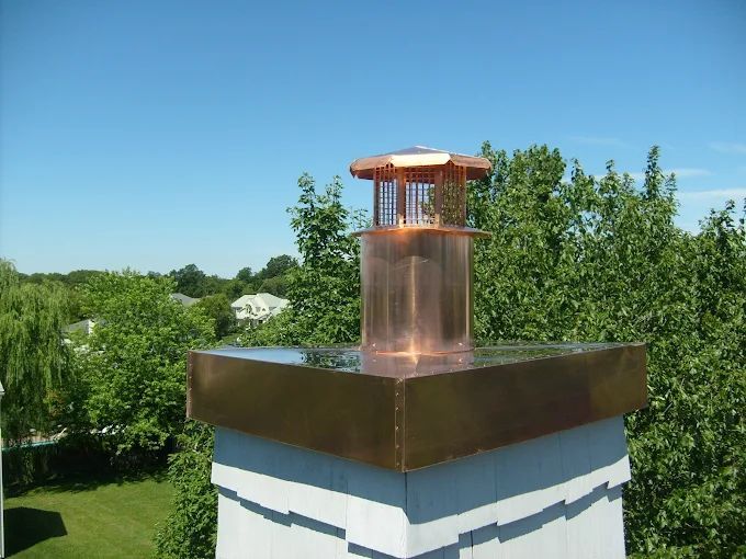 Copper chimney cap on a white rooftop against green trees and blue sky
