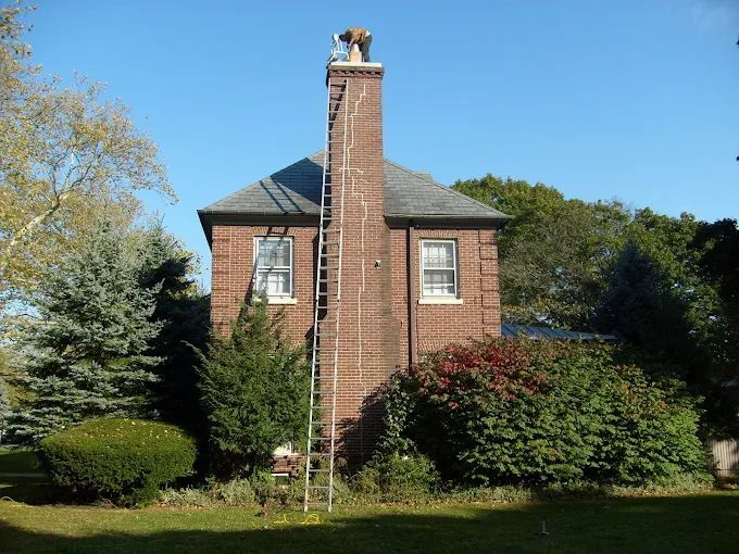 Brick house with a central chimney, surrounded by trees and shrubs under a clear blue sky