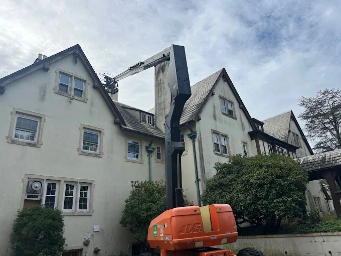 Orange cherry picker raised beside a white house with steep roofs, likely for roof repairs