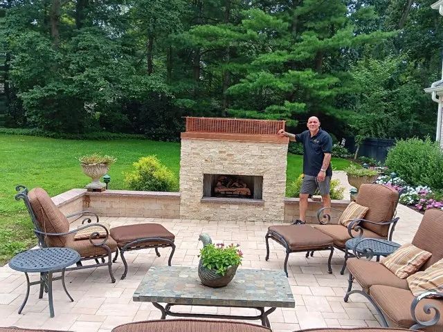 Patio with outdoor seating and brick fireplace; man stands beside it in a green backyard.