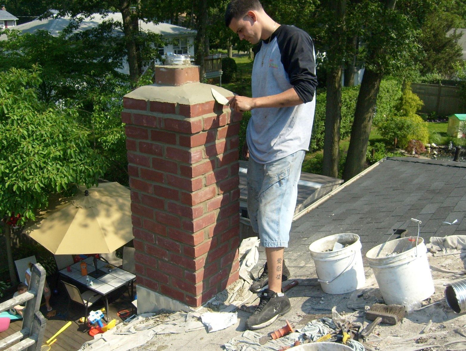 Man laying bricks on a chimney atop a roof, with buckets and tools nearby.