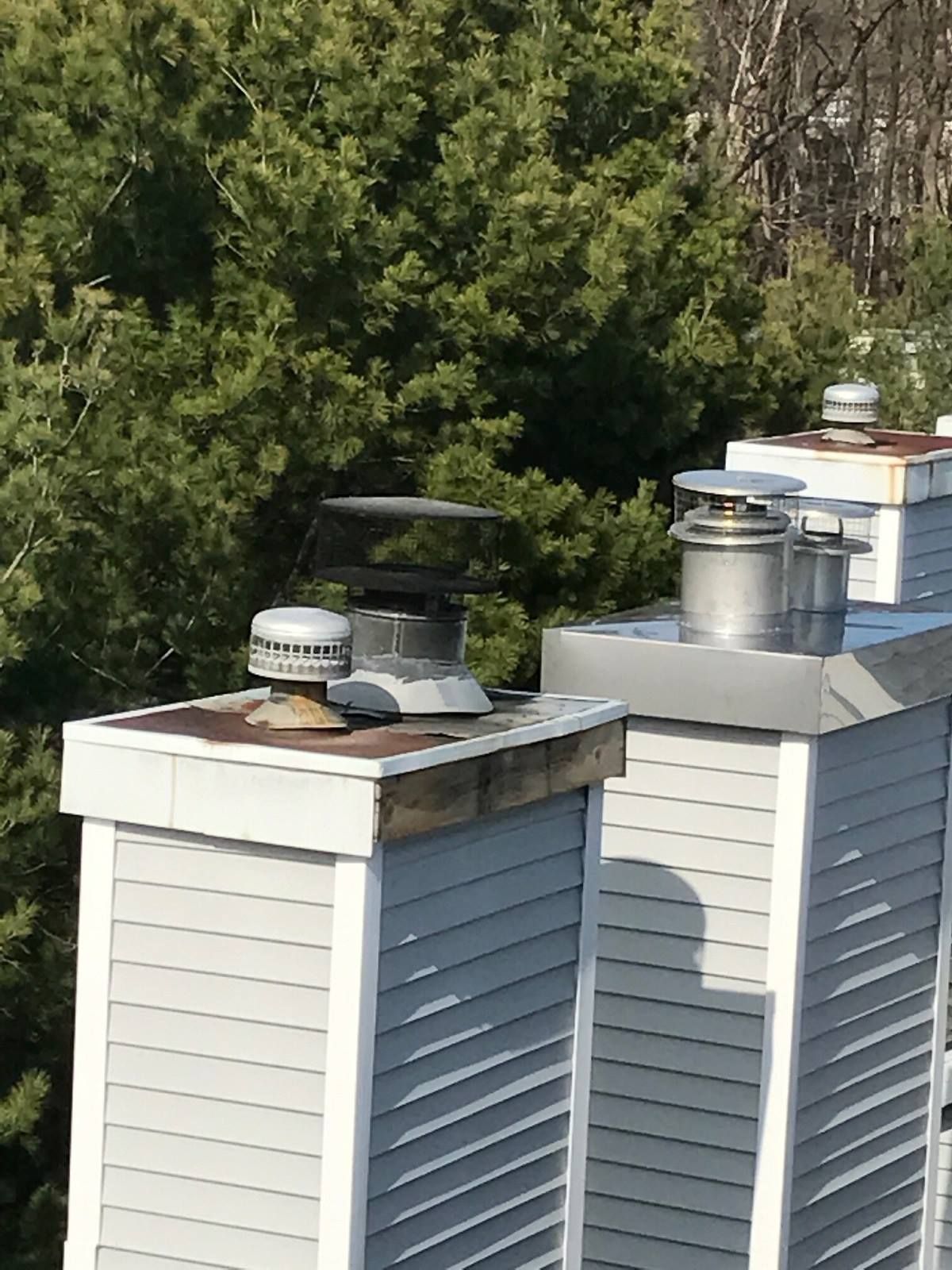 Two white roof chimneys with metal caps and vents against a backdrop of trees.